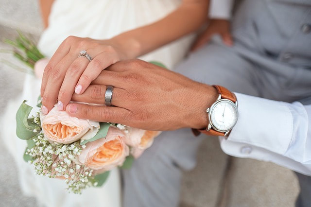 Bride and groom touching hands on top of peach colored roses to display their wedding rings.