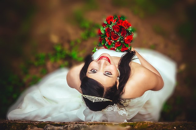 Looking down at a smiling bride from above as she poses in her wedding dress holding a bouquet of red roses.