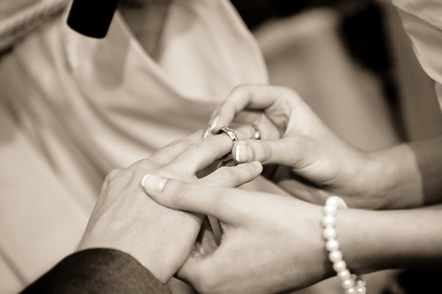 A black and white photo of a bride's hand sliding a wedding band on her new husband's finger.